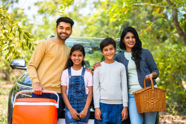 istockphoto-1483218108-612×612 (1) Happy smiling family with sibling kids standing in front of car for picnic by looking camera during holiday travel - concept of joyful lifestyle, family time and freedom.
